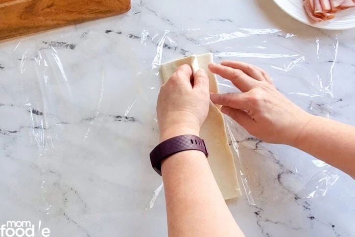 Unrolling the thawed puff pastry dough onto a sheet of plastic wrap.