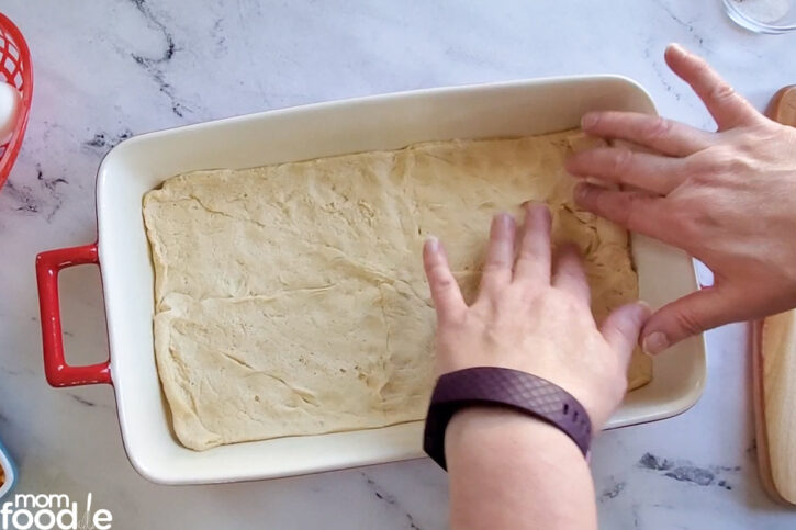 crescent roll dough placed in casserole, shows hands pinching the holes closed and pushing dough into corners.