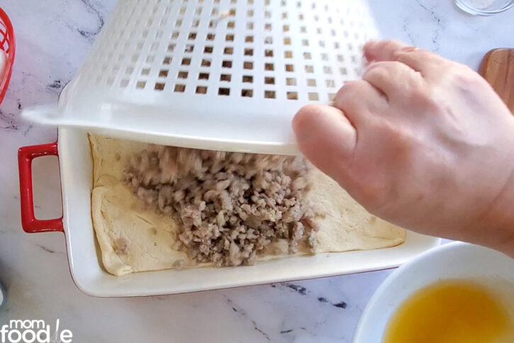 drained sausage being dumped out of colander onto the crescent dough, the drained grease in bowl in background