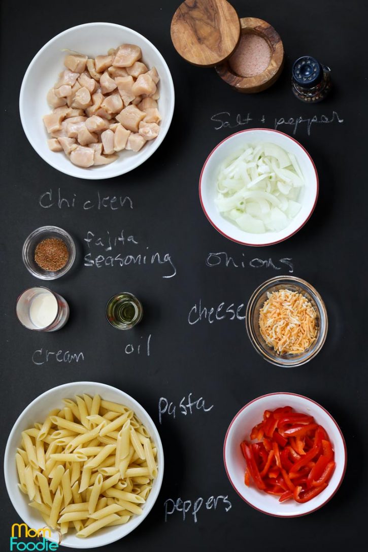 ingredients for the chicken fajita pasta recipe laid out on blackboard and labeled. chicken, cheese, oil, cream, pasta, bell peppers, fajita seasoning, onions, salt and pepper.