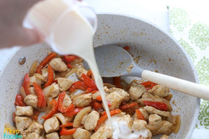 Pouring cream into the pot with chicken, red peppers and onions.
