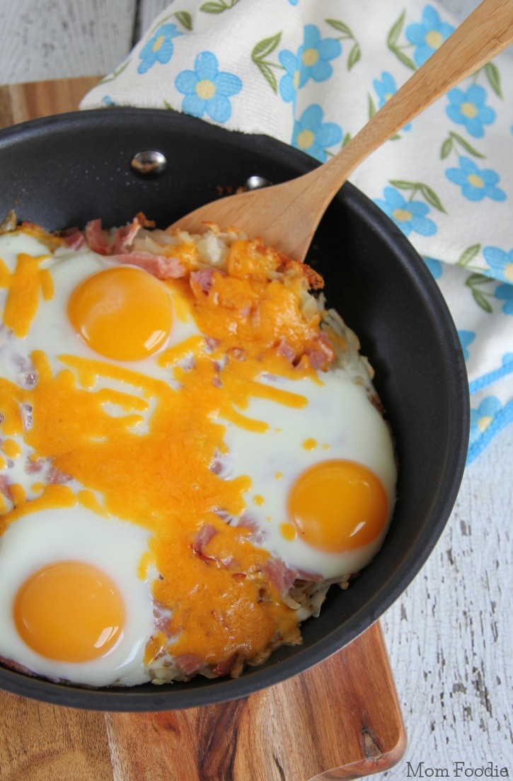 Skillet hash brown breakfast casserole on wood cutting board. the breakfast skillet has sunny side eggs on top and floral tea towel behind it.