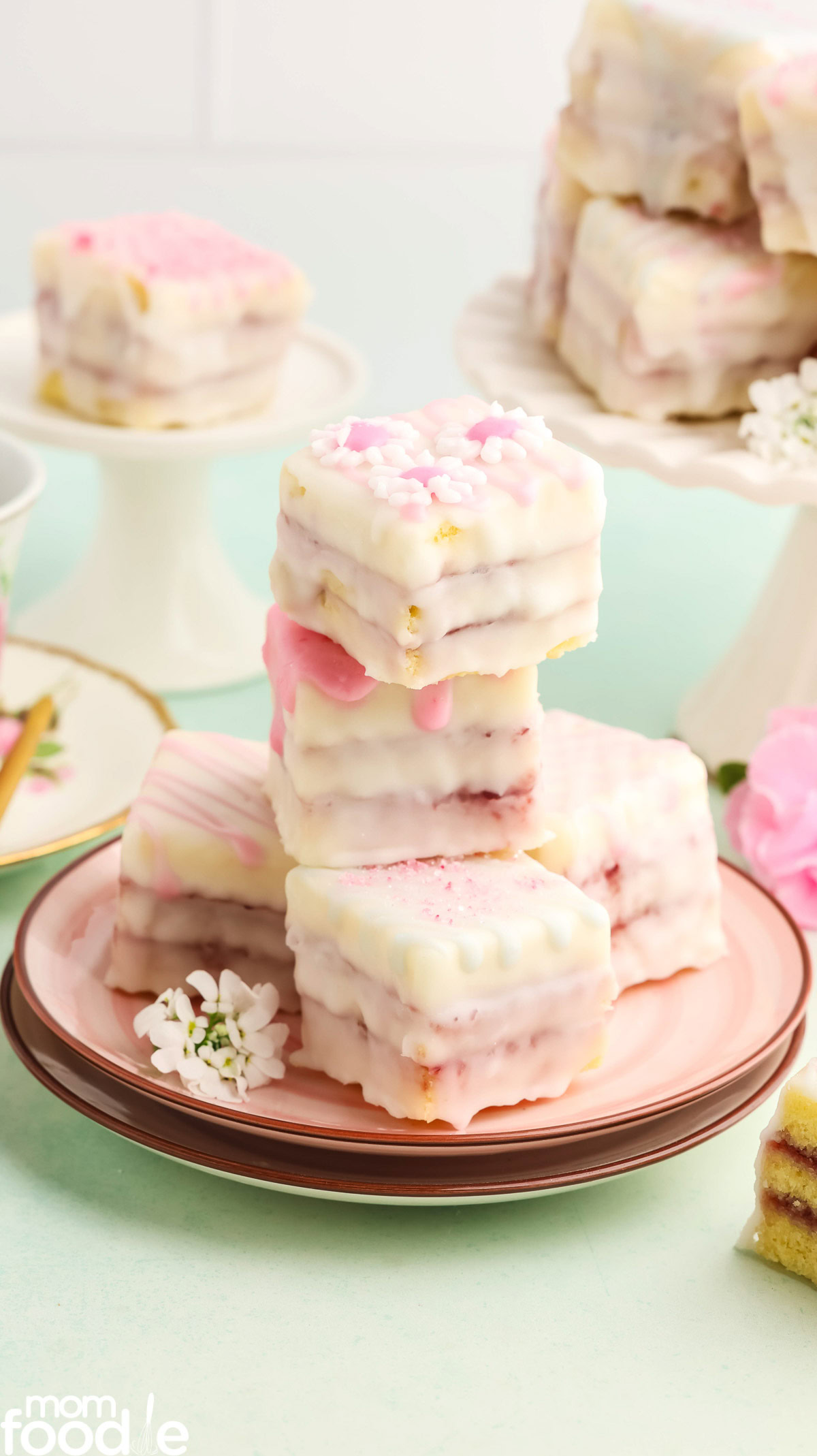 stack of petit four cakes on pink plate with more petit fours in the background