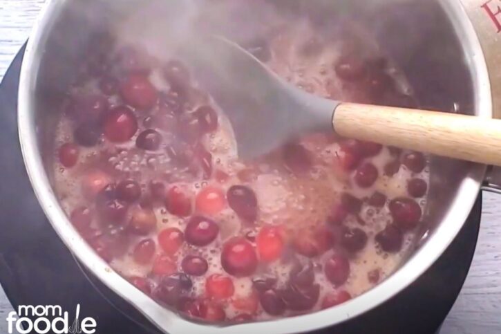 Cranberries popped in simmering sauce. Most have burst in the pot and the cranberry orange sauce is starting to thicken a bit. Shown stirring.